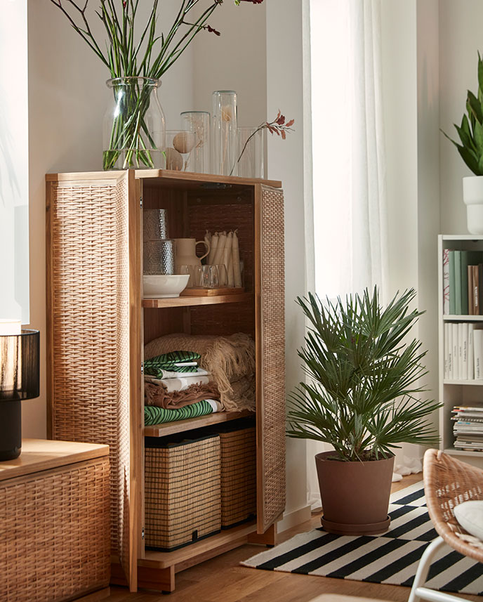 A bright living room with a FRYKSÅS cabinet made of rattan, displaying decorative items and surrounded by plants.