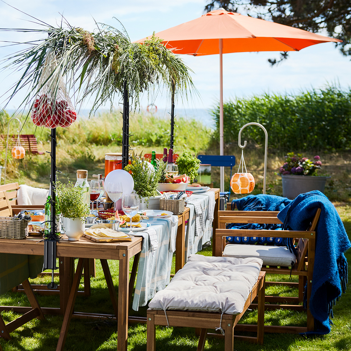 A garden setting with a light brown wooden NÄMMARÖ table with food and drinks on a sunny day. Chairs, a bench and a parasol.