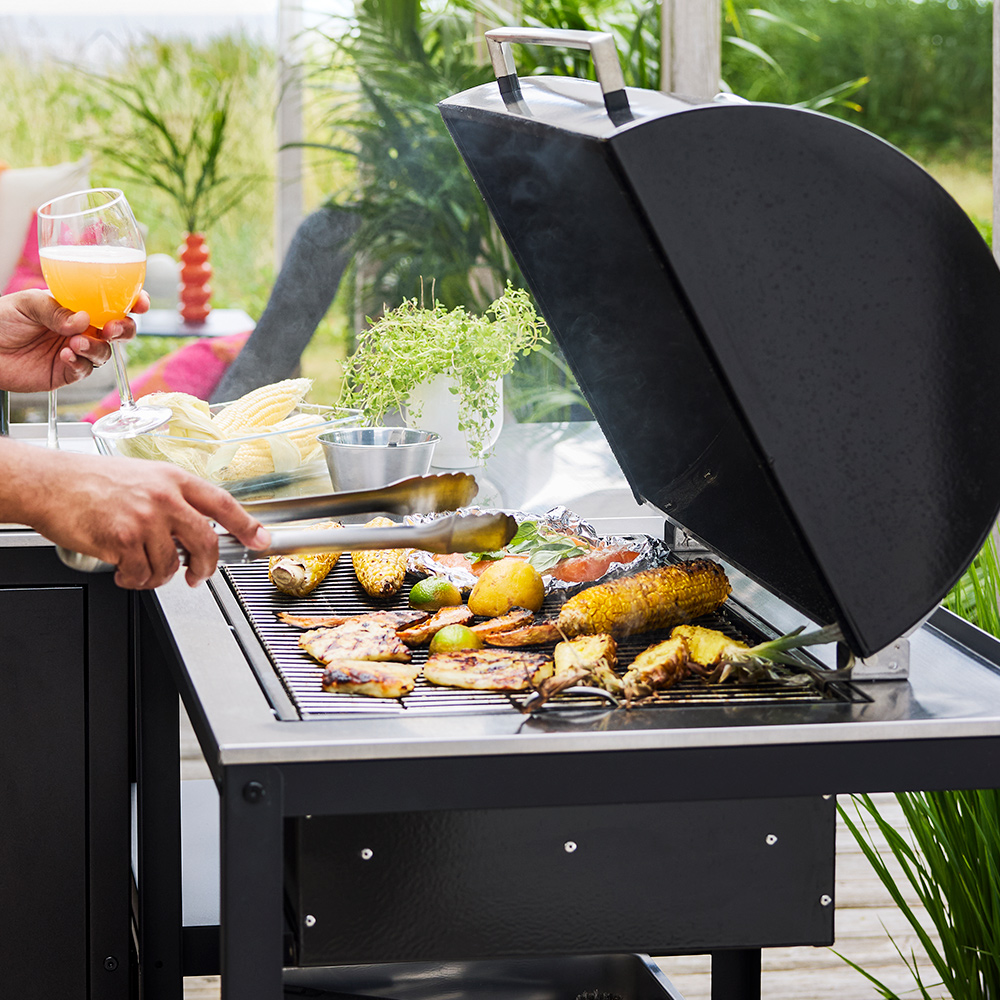 A patio with lush greenery features a black GRILLSKÄR charcoal barbecue made of black and stainless steel cooking food.