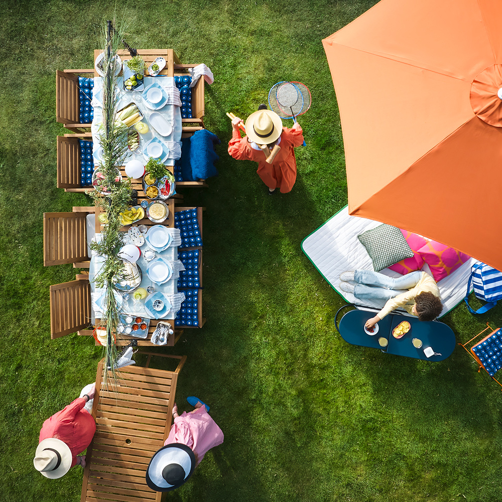 A garden with outdoor seating features a light brown stained NÄMMARÖ table surrounded by people enjoying refreshments.