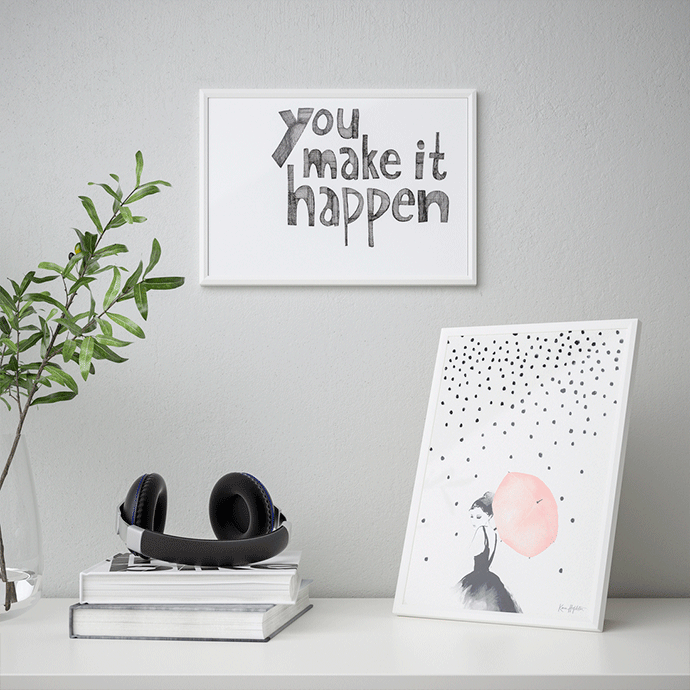 Minimalist workspace with a white framed "You make it happen" print on the wall and a leaning illustration of a girl with a pink umbrella, next to headphones and books.