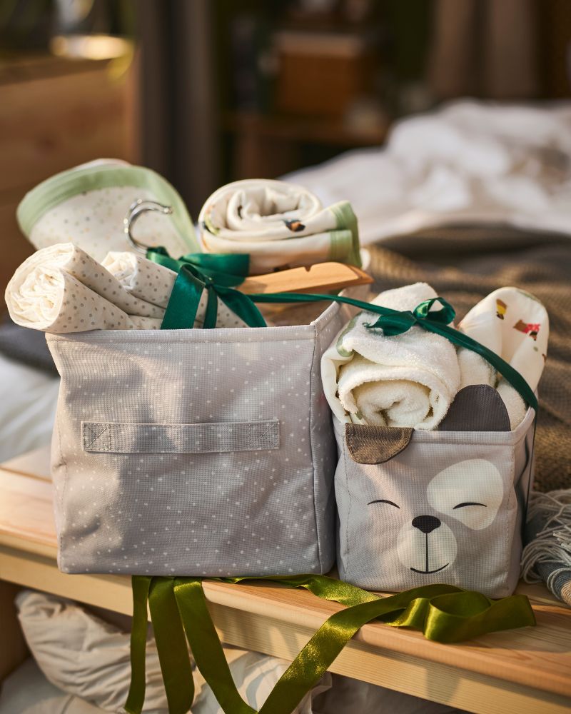 DRÖMSLOTT boxes with textiles and rolled LENAST crib sheets on a wooden bench in a warm setting.