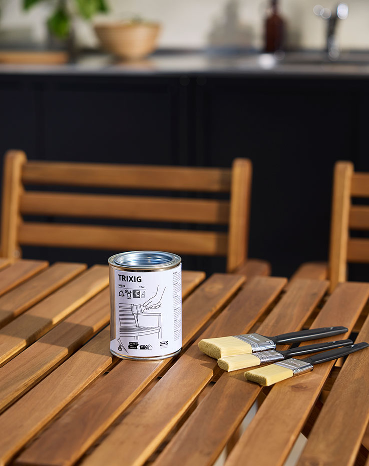 A wooden outdoor table with three brushes next to a can of TRIXIG outdoor wood stain placed on the table.
