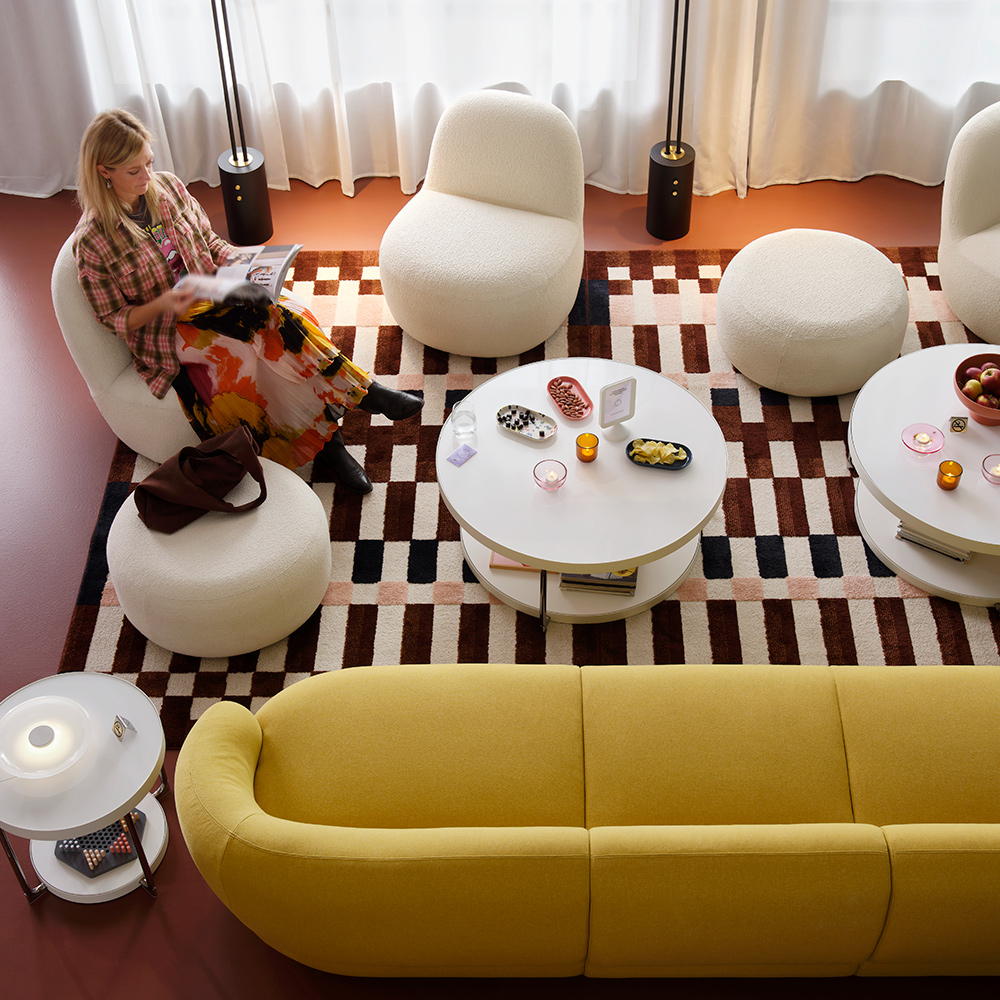 A person sits across from a round, white glass FRÖTORP coffee table holding snacks in a hotel lounge with other curvy seating.