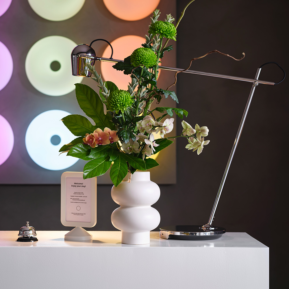 A curvy, beige stoneware PÄRLHÄGG vase holds fresh flowers on a white hotel reception desk, by a lamp, a bell and signage.