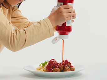 Una persona vierte ketchup desde un bote rojo sobre un plato de albóndigas vegetales, añadiendo el toque clásico sueco.