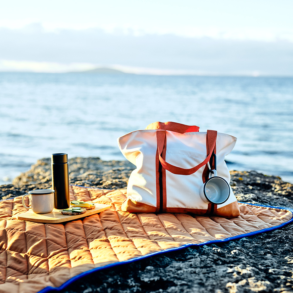 A SOLUPPGÅNG bag, two mugs and a chopping board are on a SOLUPPGÅNG picnic blanket on a rocky cliff by the sea.