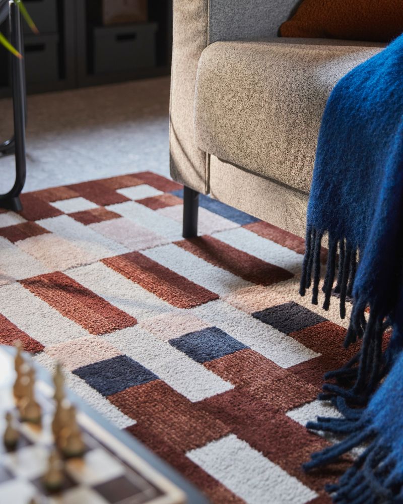 A living room features a multicoloured MARKBLÅMME low pile rug with a check pattern under a sofa and next to a chess set.