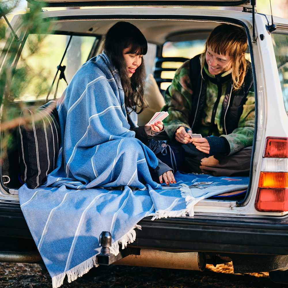 A couple is smiling and playing cards in the back of a car; one of them has a SOLUPPGÅNG throw wrapped around her shoulders.