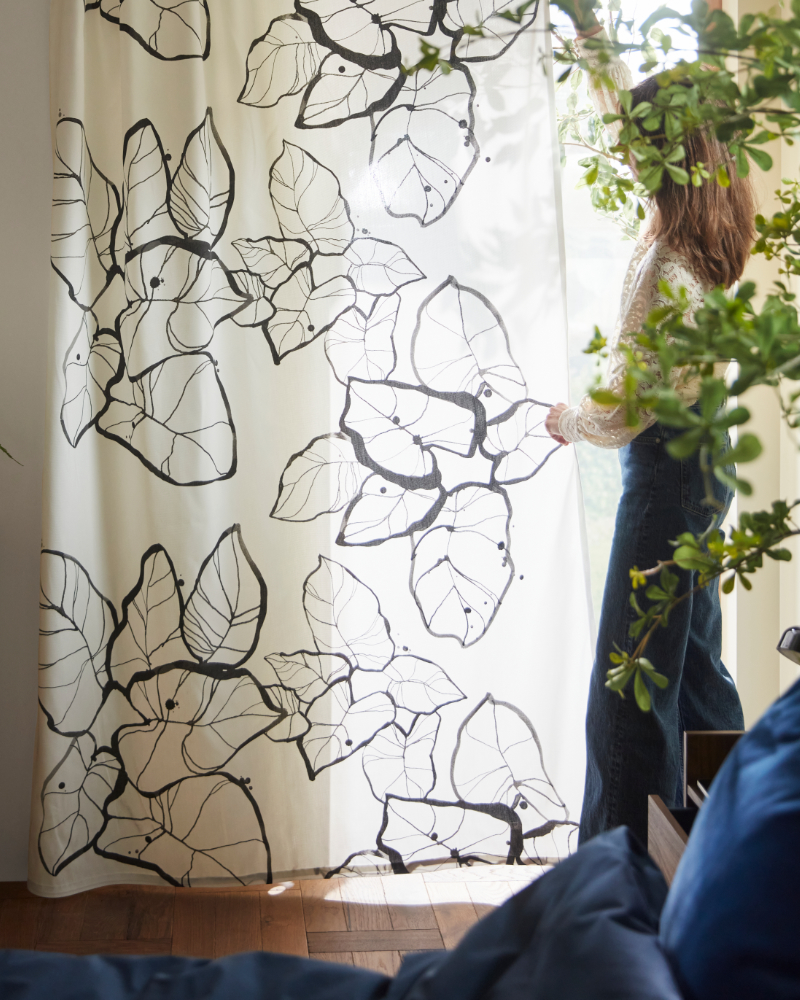 A living room features white and black PELARKÖRSBÄR curtains with a floral pattern, hanging beside a large window.