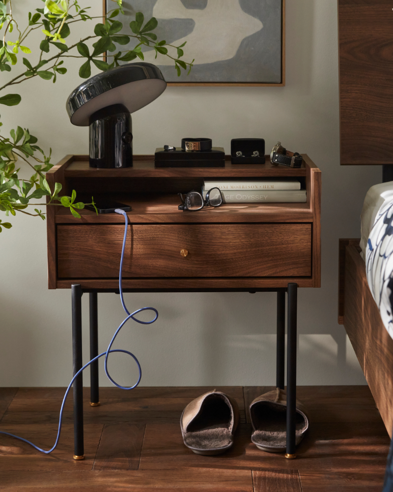 A bedroom featuring a brown RÅDMANSÖ bedside table with a walnut effect on legs, beside a bed and a potted plant.