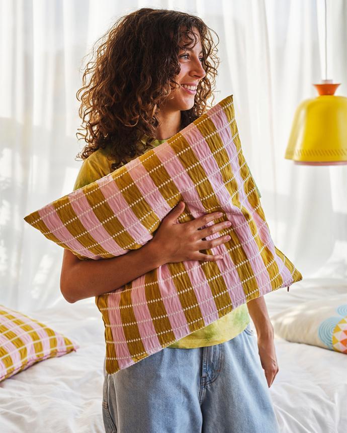 Smiling woman holding a checkered decorative cushion in a bright room.