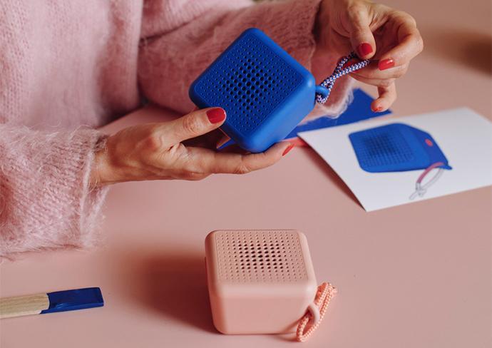 Hands holding a blue portable speaker on a table with other small devices.