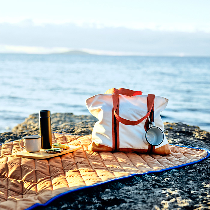 Coastal picnic scene with a tote bag, quilted blanket, and accessories on rocks by the sea.