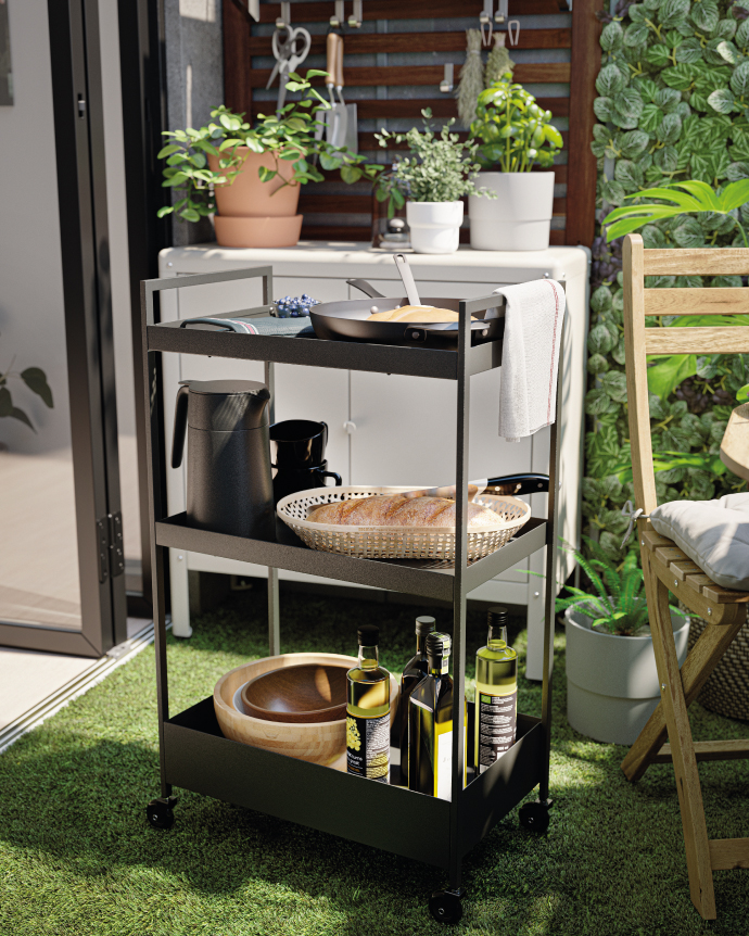 Black three-tier serving cart on a patio with plants, holding a pan, kettle, bread, oil bottles, and kitchen utensils.