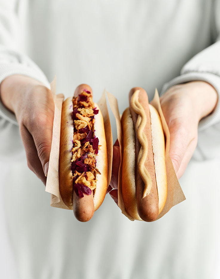 Close up of a person’s hands holding two hot dogs in paper sleeves; one has red cabbage and crispy onions, the other has a smooth mustard drizzle. 