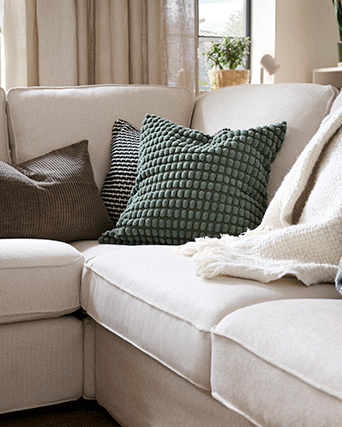 A green textured cushion among others on a light beige sectional sofa near a window.
