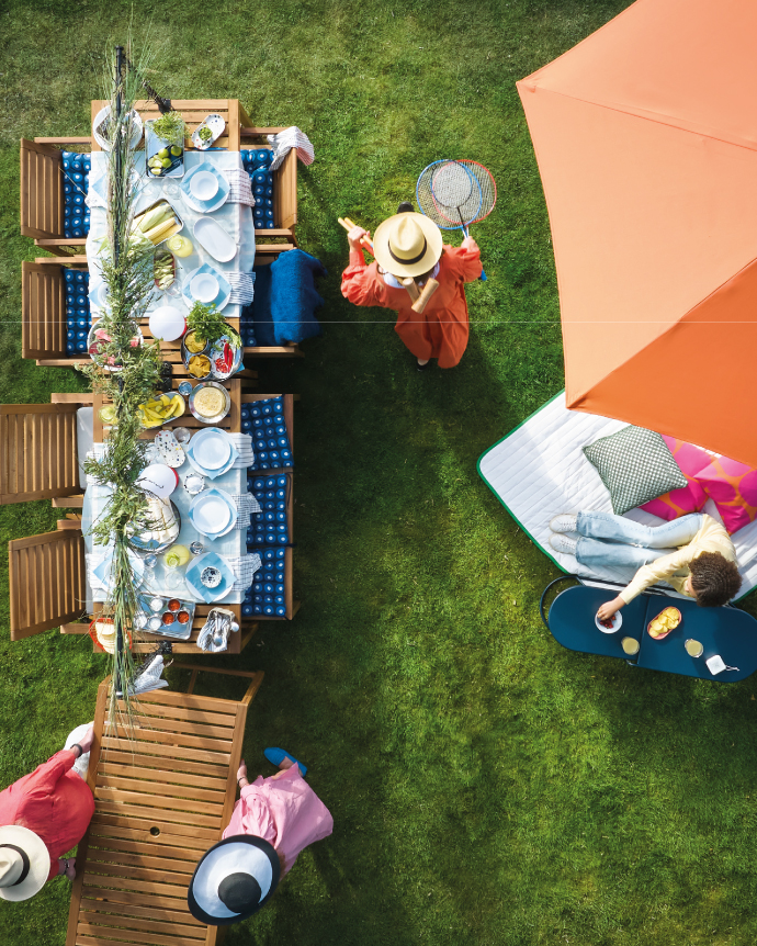 Overhead view of an outdoor decorated table with plates, fruit, and drinks, while several people enjoy the garden; one person holds a racket and another relaxes under an orange umbrella.