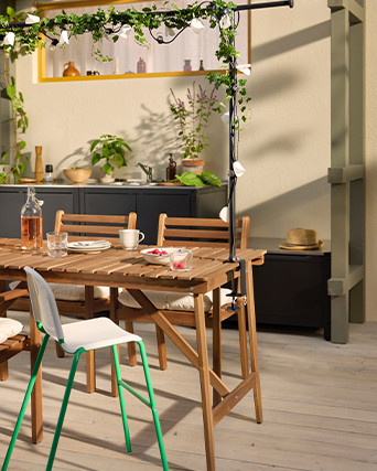 Mesa de comedor exterior de madera con bebidas, aperitivos y un sombrero de paja, bajo una guirnalda decorativa de hiedra y luz solar cálida.