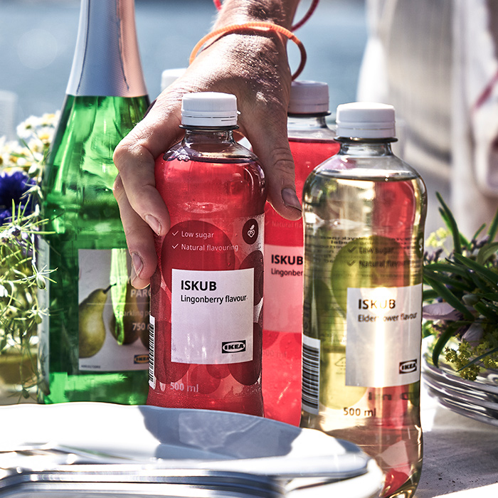 Outdoor table by the sea with bottles of ISKUB lingonberry drink and pear cider. A hand reaches for a red bottle near stacked white plates.