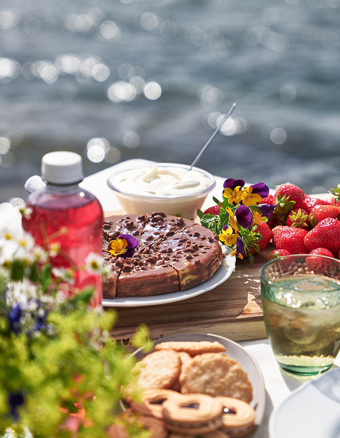 A sunny picnic spread with fresh strawberries on a wooden board, chocolate cake, biscuits, and a cold drink, with the sea blurred in the background.