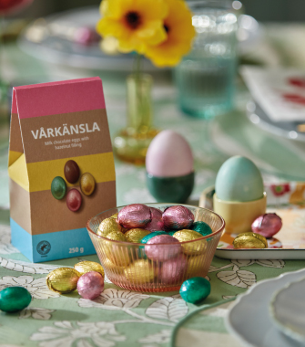 Una mesa festiva con una bolsa de papel que dice "VÅRKÄNSLA", un cuenco acanalado lleno de huevos de chocolate con papel de aluminio multicolor, dos huevos pastel en tazas y flores amarillas en un jarrón, todo sobre un mantel verde con diseño.