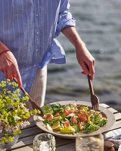A close-up photograph of a person in a blue striped shirt serving a salad with salmon pieces from a white plate using wooden servers, at an outdoor wooden table by the water.