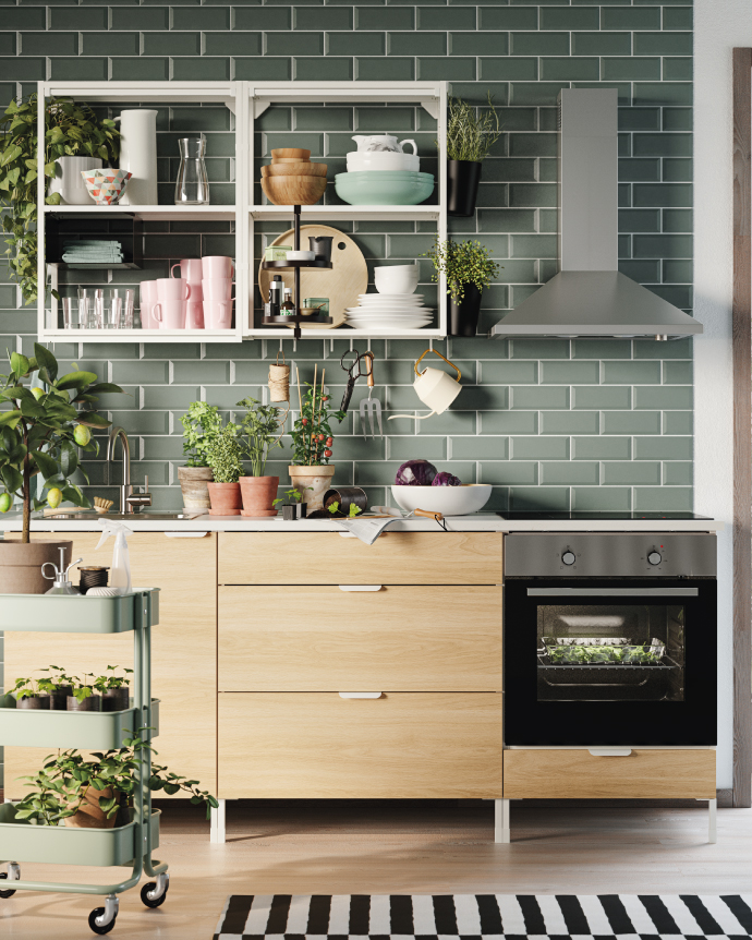 Bright kitchen with green subway tiles, open shelves displaying dishes and cups, potted plants, and a built-in oven in light wood cabinetry.