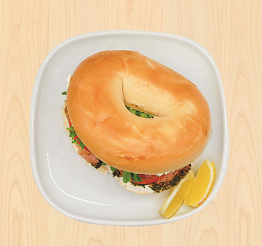 Bagel sandwich filled with salmon, fresh vegetables, and seeds, served on a white plate with lemon wedges on the side, on a light wooden table.