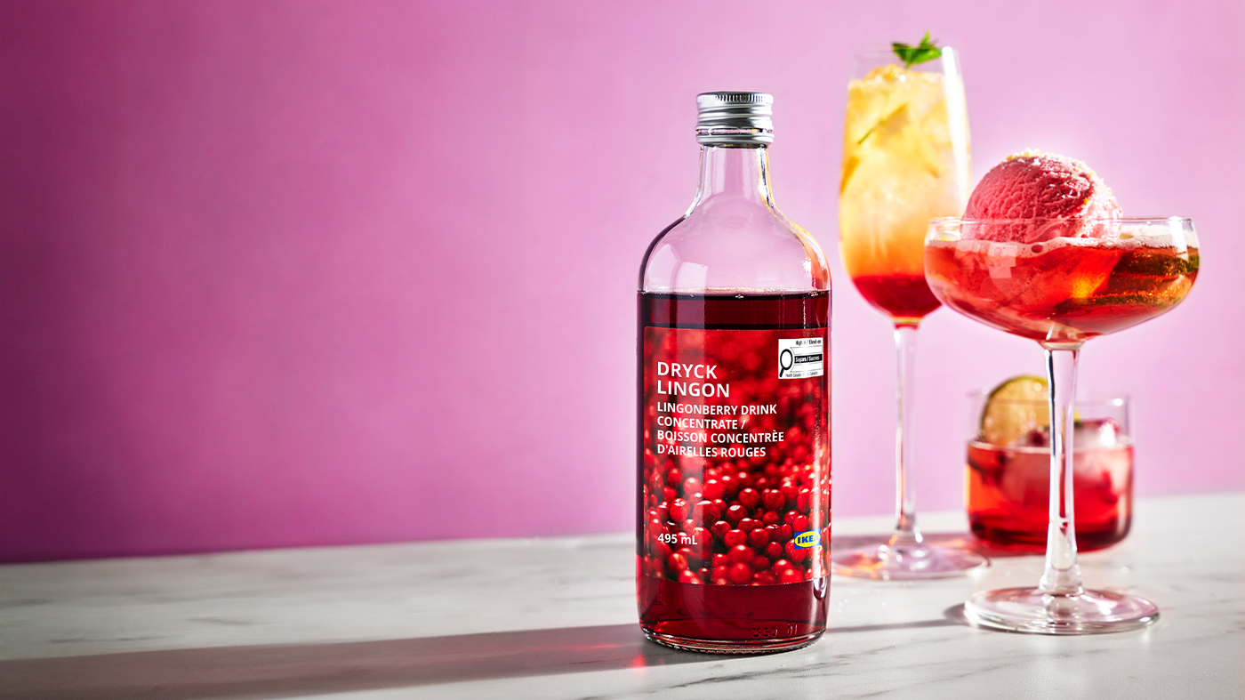 Bottle of lingonberry concentrate with red cocktails in elegant glasses against a pink background.
