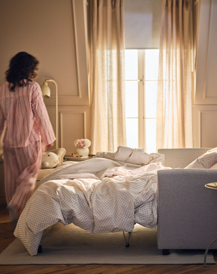 A person stands beside a light beige EKHOLMA 2-seat sofa-bed which is configured as a bed and has BANDKNIPPE bed linen.