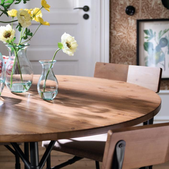 Close-up of a round wooden dining table with white and yellow flowers in glass vases under natural light.