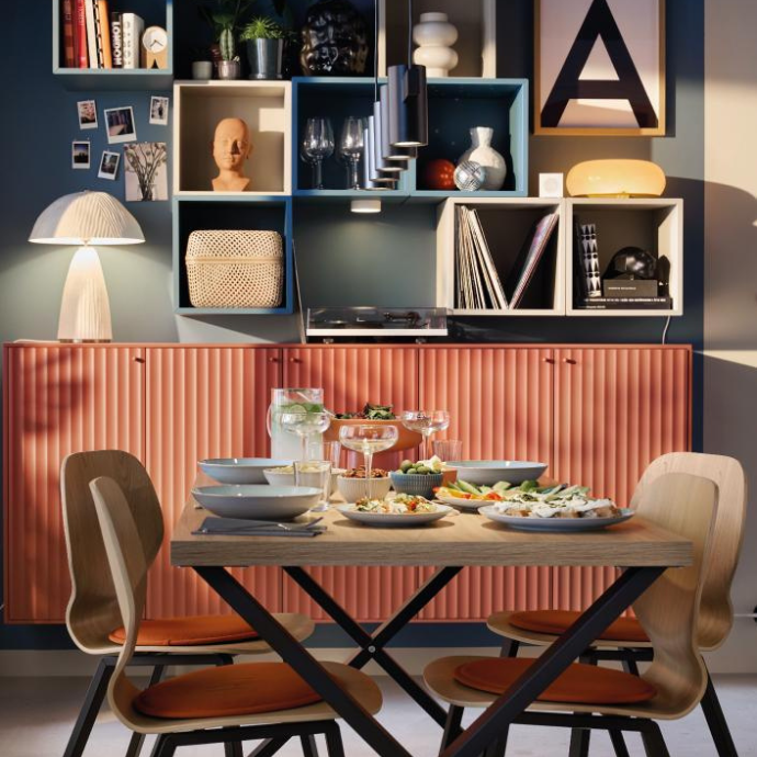 Modern dining room with a wooden table, orange-cushioned chairs, and a colorful modular bookshelf in the background.