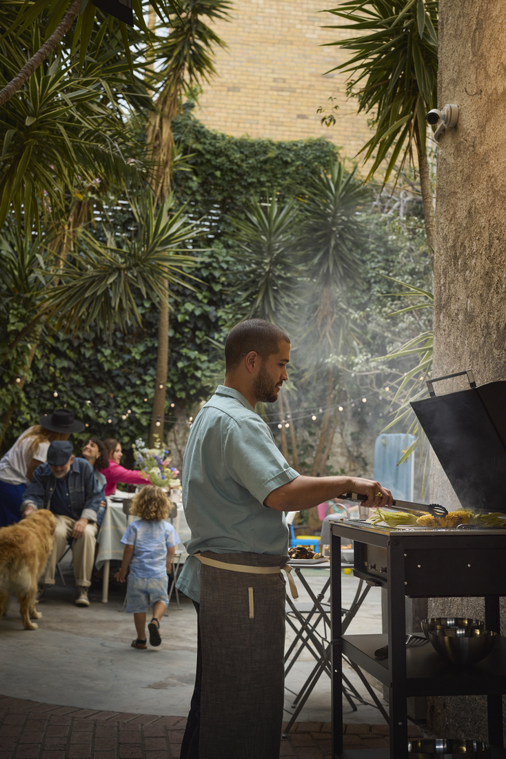 A man in an apron grills corn on a barbecue during a family gathering in a patio with palm trees.
