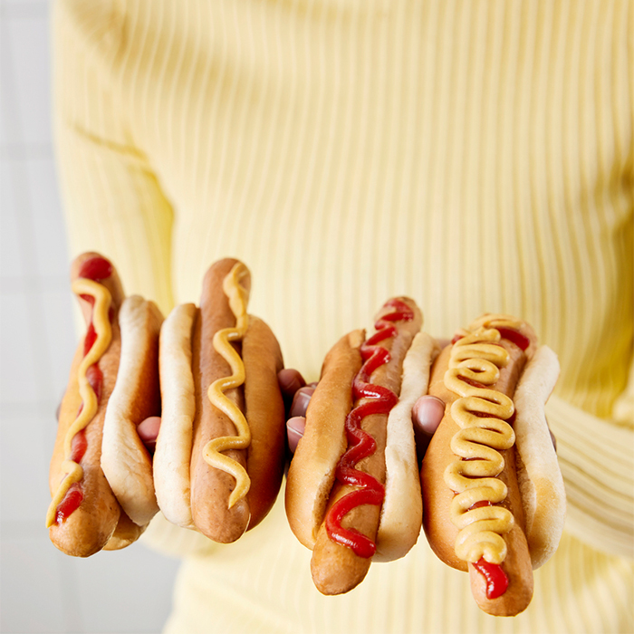Close-up of a person in a yellow sweater holding four hot dogs with various ketchup and mustard toppings in a bright kitchen.