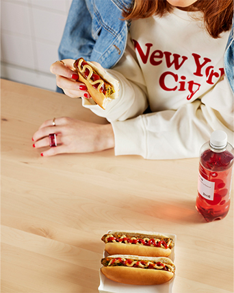 A person in a "New York City" sweatshirt holds a bitten hot dog at a table with more hot dogs and a red drink.