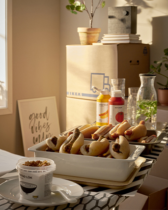 Multiple hot dogs and fried onions on a makeshift table among IKEA moving boxes and condiments in warm sunlight.