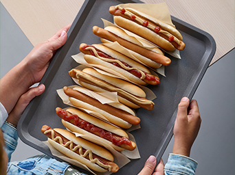 Top-down view of a person in a denim jacket holding a grey tray with seven hot dogs featuring various toppings.