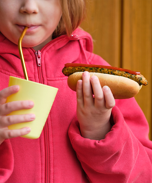 Close-up of a girl in a pink hoodie drinking from a yellow cup with a straw while holding a veggie hot dog topped with ketchup.