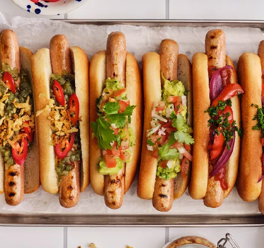 Top-down view of a row of gourmet hot dogs with various toppings including guacamole, pico de gallo, red onions, and chilies on a parchment-lined tray.
