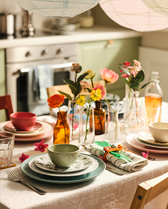 Mesa de comedor decorada con vajilla colorida, flores frescas en jarrones de vidrio y mantelería de lino en una cocina acogedora.