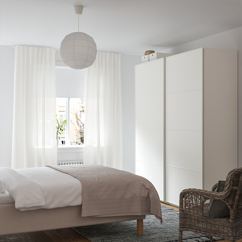 Minimalist neutral-toned bedroom featuring a large white wardrobe, beige bed runner, sheer curtains, and a wicker armchair.
