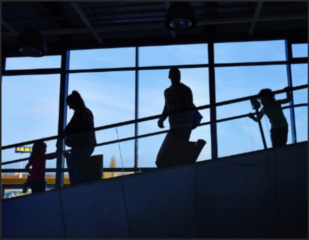 Silhouettes of several people walking along an indoor walkway in front of large, backlit windows.