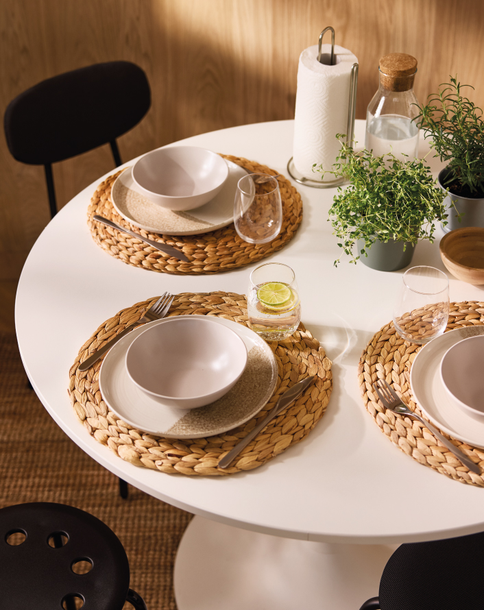 White round table set with plates, cutlery, glasses, and decorative plants in a warm setting.