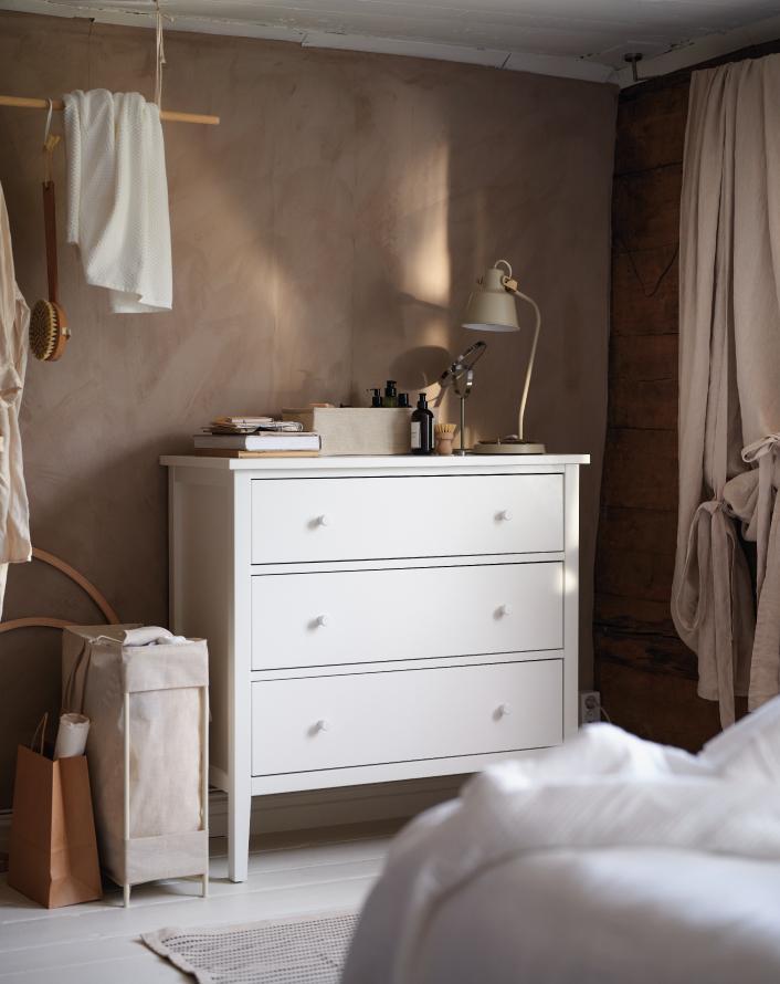 A white three-drawer dresser in a rustic room with textured walls, a desk lamp, and a laundry bag nearby.
