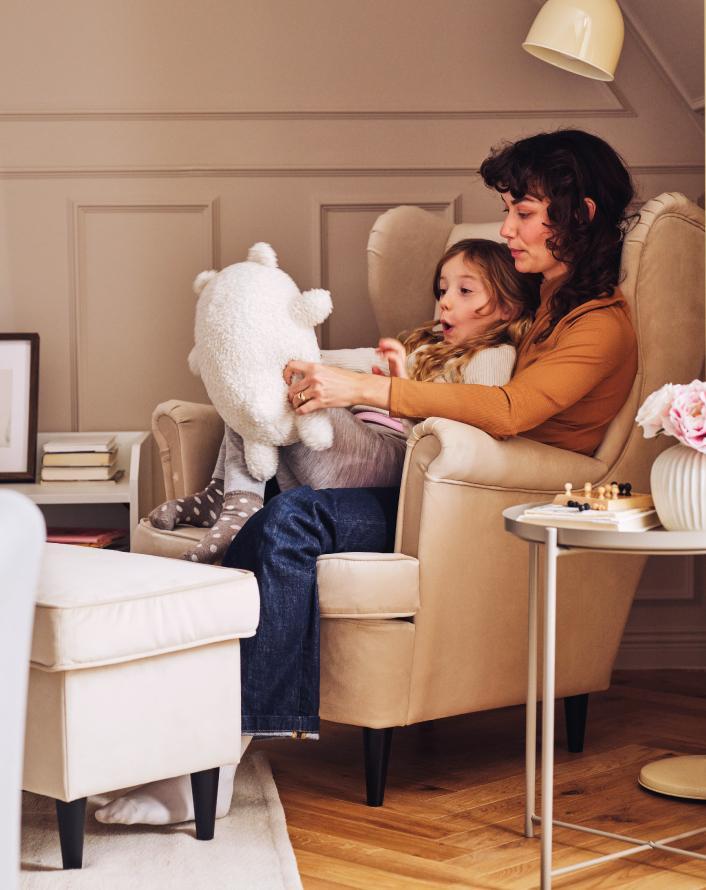 A mother and daughter sitting together in a beige wingback armchair, holding a large white plush toy.