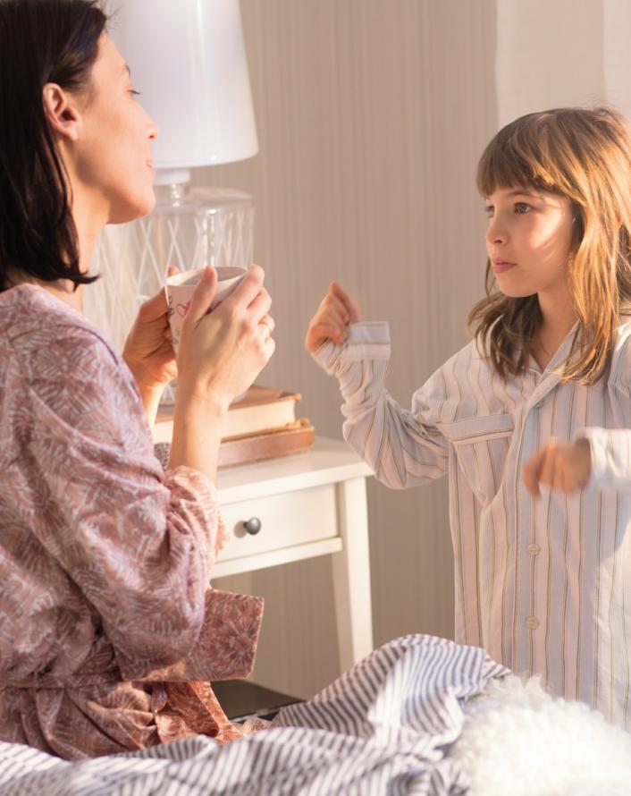 A woman in a patterned robe holding a mug while talking to a young girl in striped pajamas in a cozy bedroom.