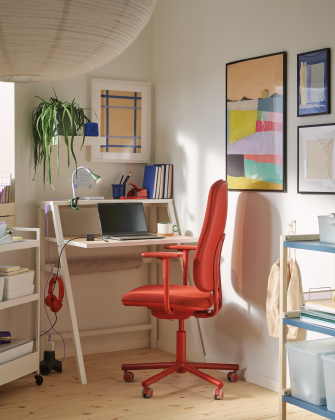 Un rincón de trabajo con un escritorio blanco pequeño, una silla de oficina roja y cuadros coloridos en la pared.