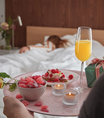 Primer plano de una bandeja rosa sostenida hacia una persona durmiendo, con dulces de corazón, un pastelito, mimosas y velas encendidas.