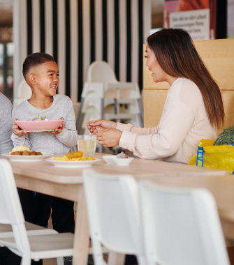 Vista de ángulo bajo de una madre sonriendo a su hijo pequeño, quien sostiene un bol de comida en una mesa de comedor con varios platos.